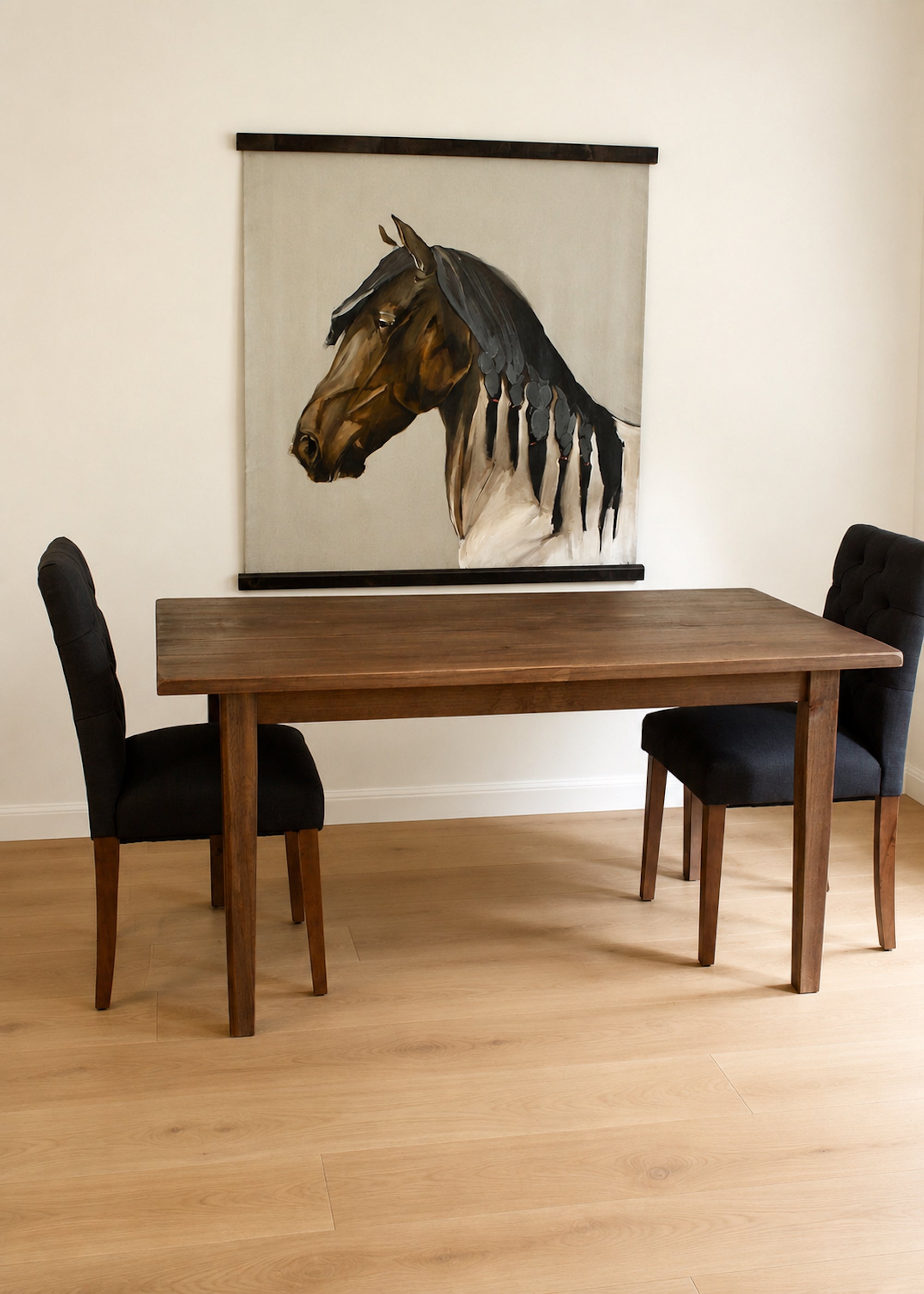 A large wall scroll painting of a horse head is displayed above a solid wooden dining table with two black chairs in a bright room with light wood floors.