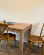 A rectangular reclaimed wood parsons dining table with a straight leg design, displayed in a home setting with plates and glasses on top. With two wood chairs