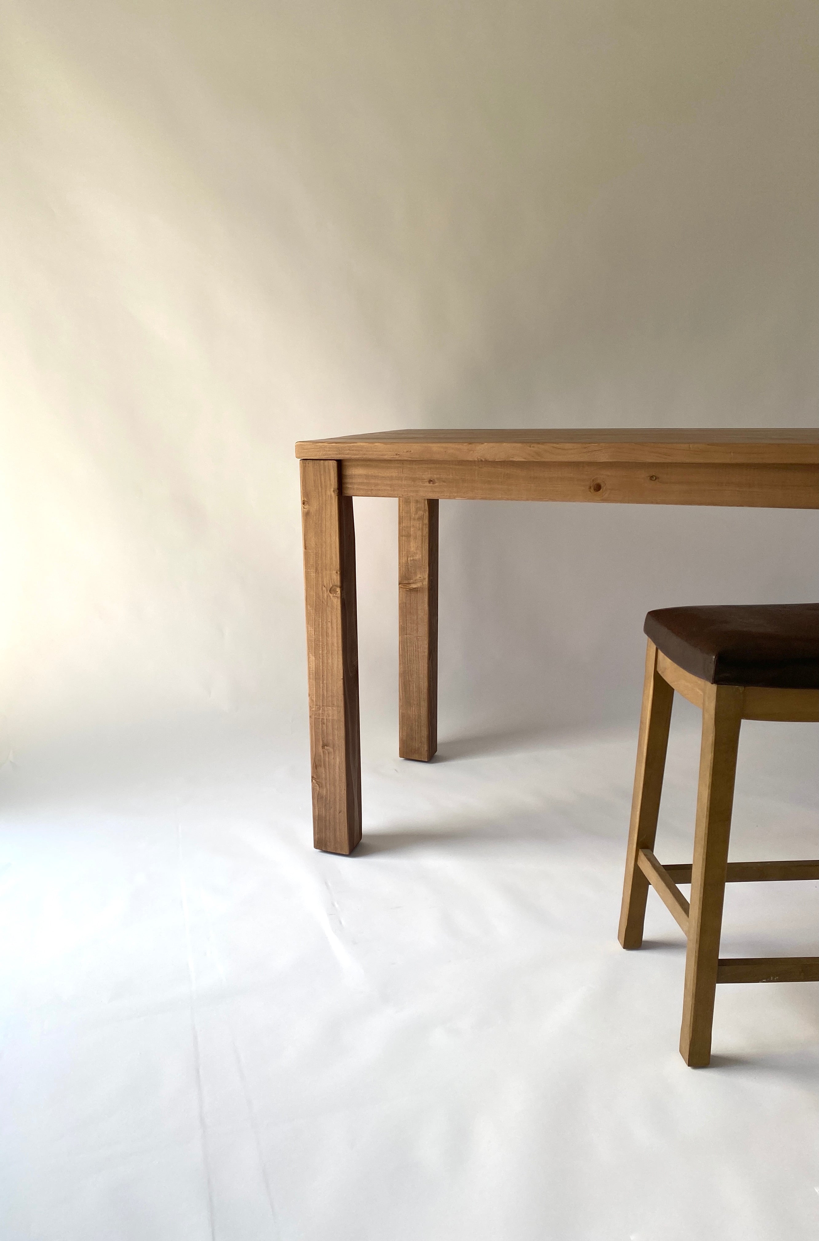 A Parsons kitchen island table made of solid wood with a straight leg design, displayed alongside a wooden stool. The table has a medium brown wood finish and is placed against a neutral background.